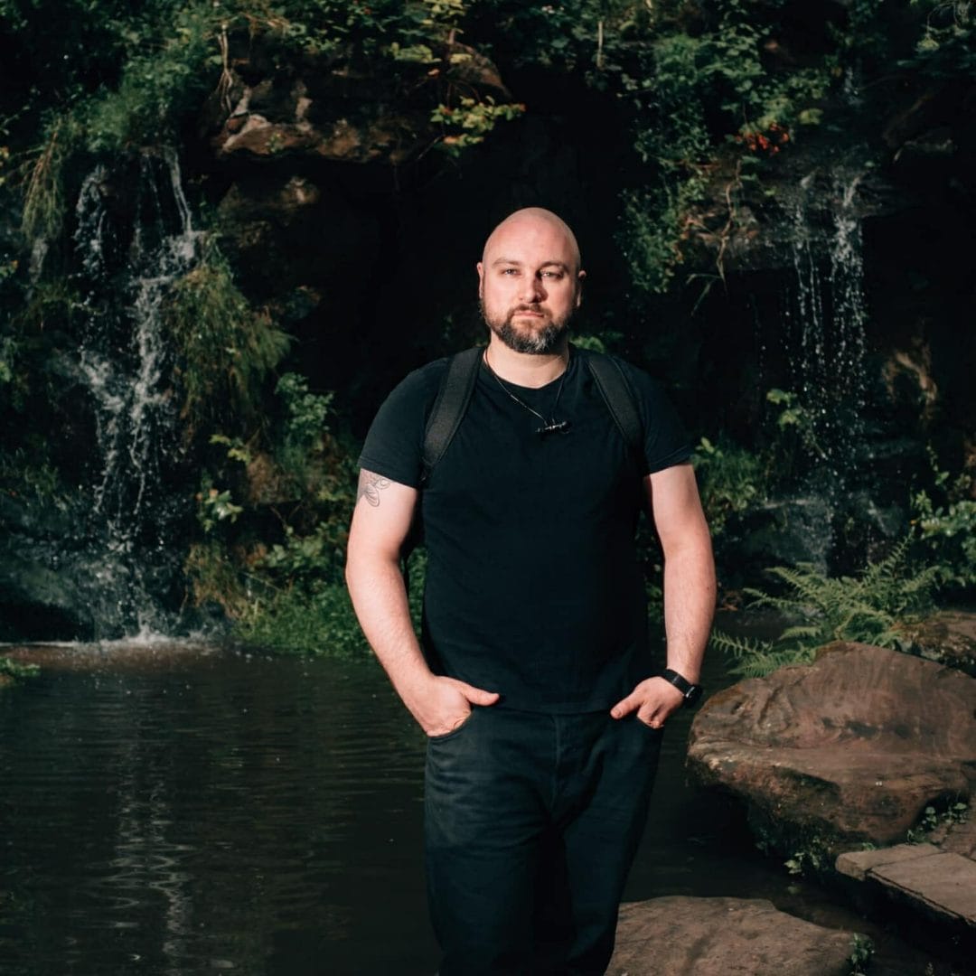 a wedding photographer standing in front of a waterfall with his hands in his pockets