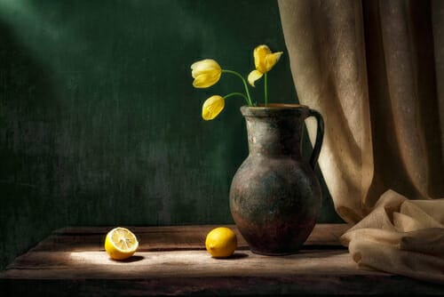 a jug sat on a wooden table with yellow flowers inside. Lemons are on the table