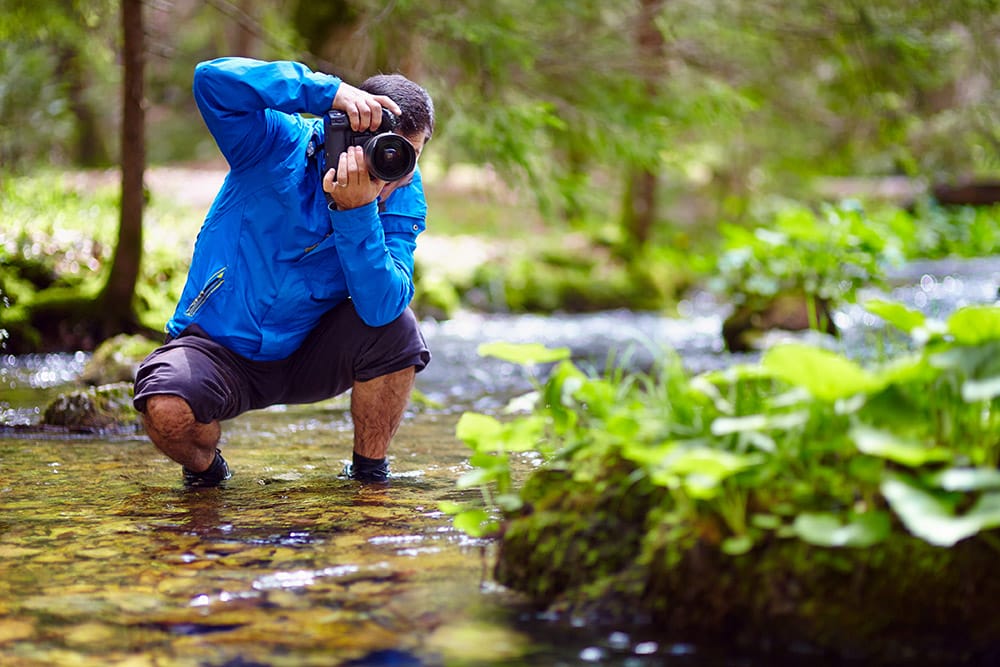Photographer crouched in woodland river in a blue jacket