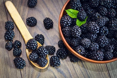 Ripe blackberries with leaves in a clay bowl on a light wooden background. Flat lay, top view. Photo of blackberry in clay bowl on wooden table. High resolution product.