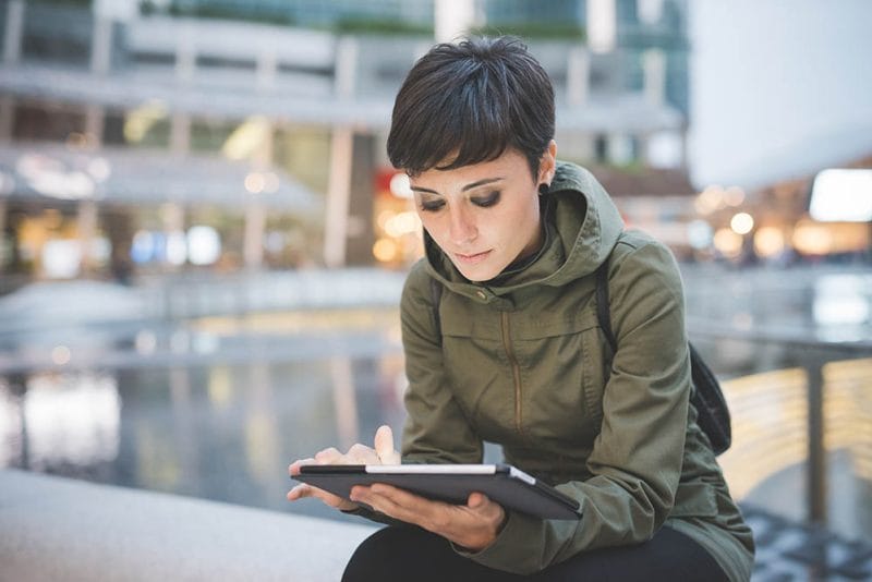 young handsome caucasian brown straight hair woman sitting in city dusk, holding tablet, looking downward screen, face illuminated by screen light - technology, social network, communication concept