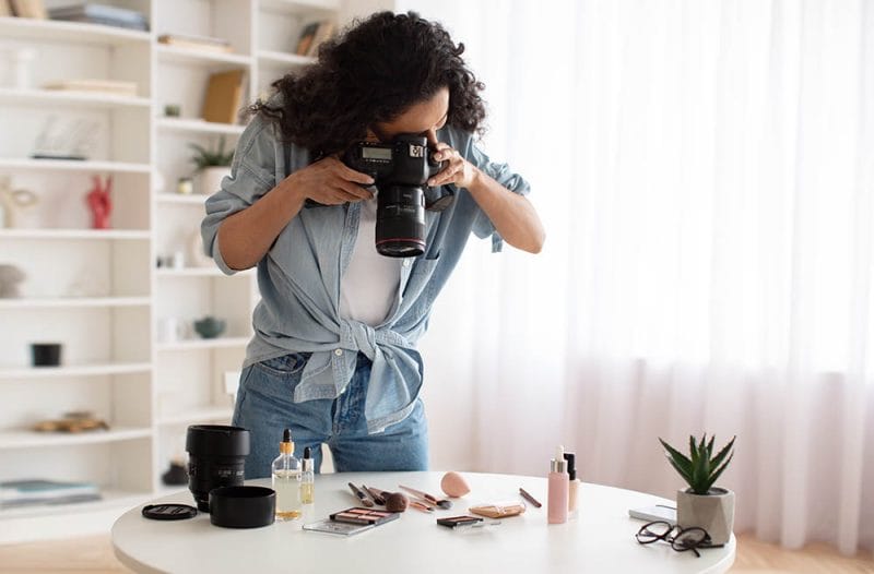 Professional Photography. Blogger Woman With Camera Taking Photos Of Makeup And Skincare Products Standing Near Desk At Home. Beauty Blogging Career Concept