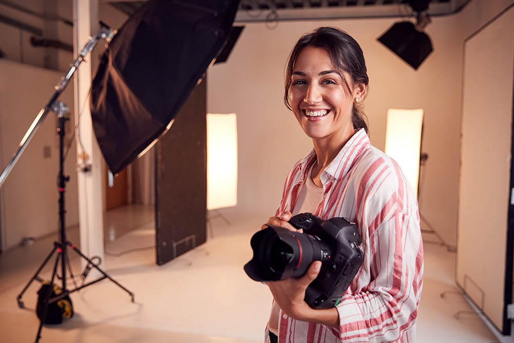 Portrait Of Smiling Female Photographer Standing In Studio With Camera And Lighting Equipment