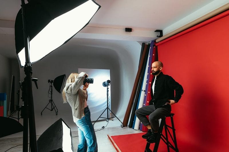 Woman photographer making business portraits for handsome bearded man on red background in photo studio. Work of a photographer. Backstage photo