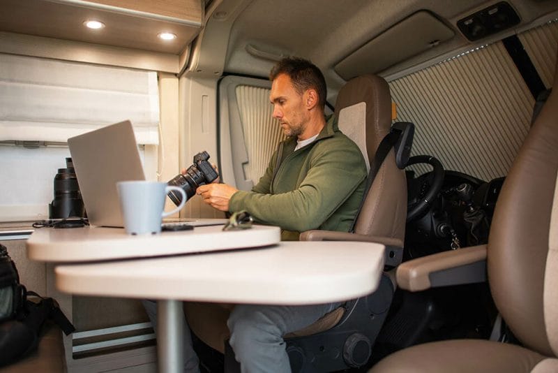 Caucasian Man in His 40s Sitting in a Modern Camper Van In Front of a Laptop with a Camera in His Hands, Transferring Recently Taken Landscape Photos to His Computer. Travel Theme.