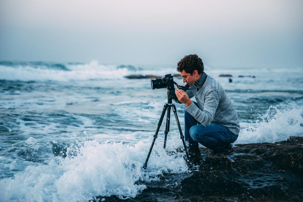 Young handsome photographer with curly hair, a gray sweater and jeans with a tripod and a camera on the beach, drenched in waves. Photography in Israel. Copy space. Shtorm on Mediterranean Sea