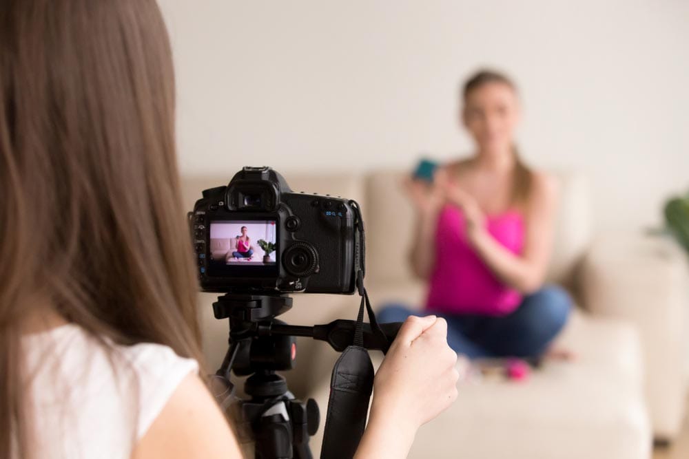 Female photographer looking at contemporary camera display, adjusting angle, preparing to take photo or video of girl sitting on sofa. Commercial product shoot, fashion photography, modeling concept.