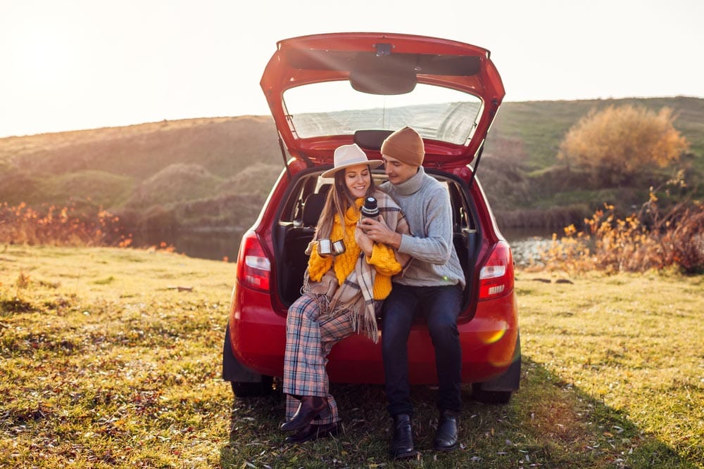 Young couple in love relaxing in car trunk by autumn lake drinking tea from vacuum flask. Man and woman travel by auto. Young man and woman enjoy fall landscape