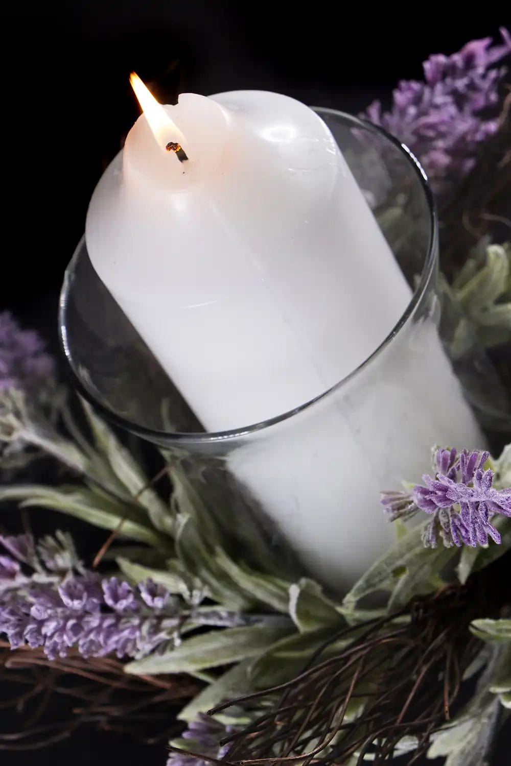 A lit white candle in a glass vase with lavender flowers surrounding