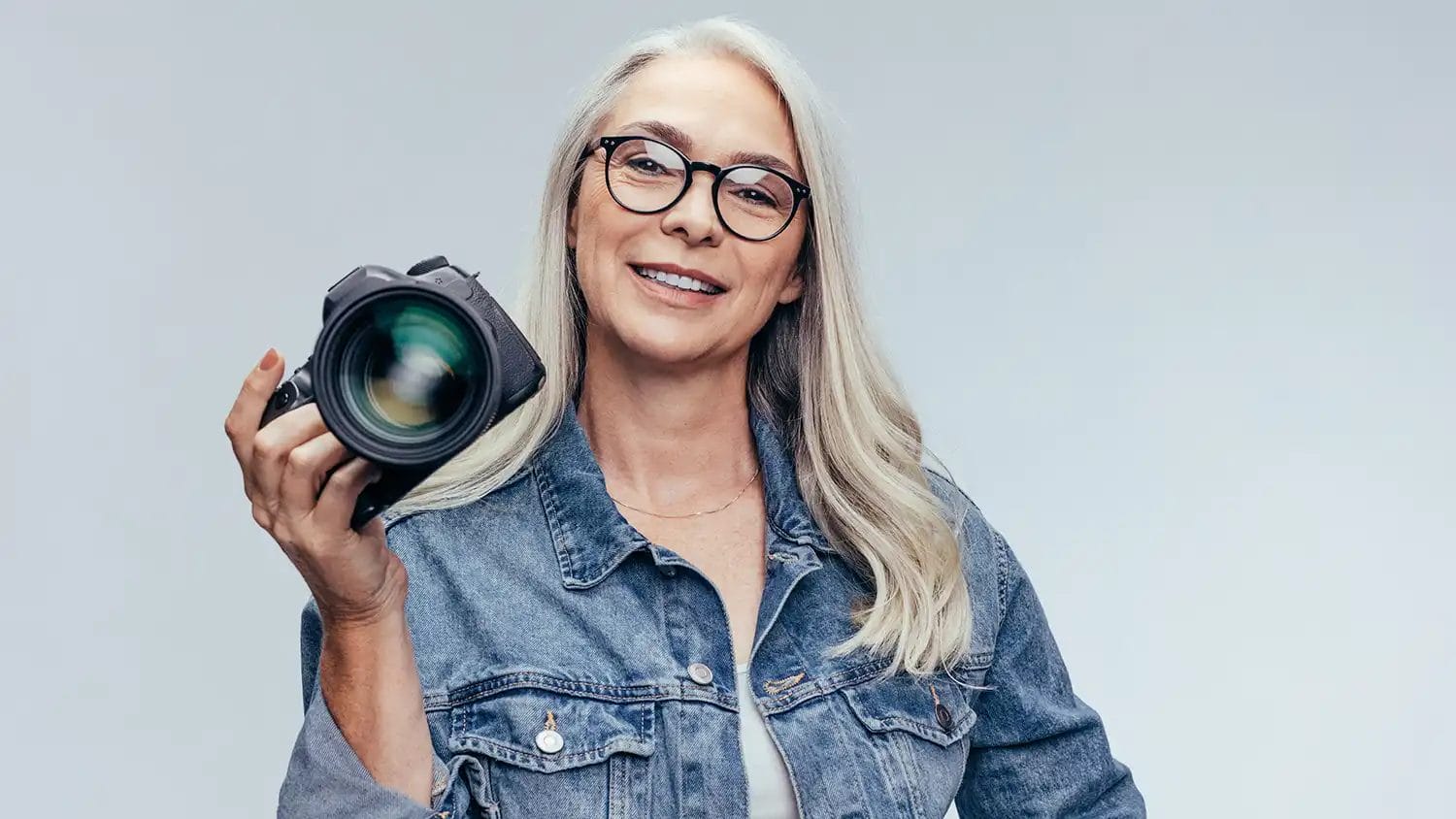 a lady, wearing glasses, with white hair holding a camera wearing a denim jacket