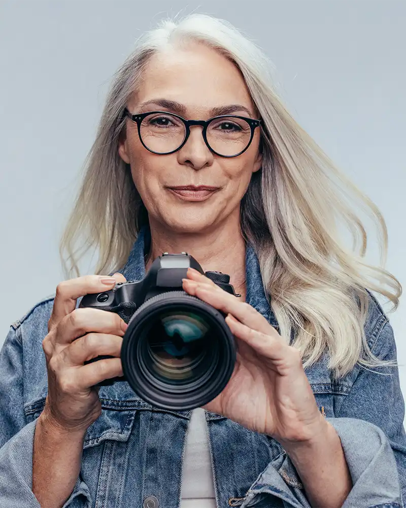 a senior lady with long white hair wearing glasses and denim jacket holding a digital camera