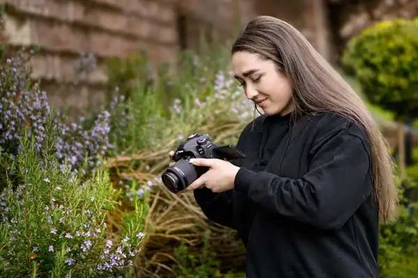 a young female photographer in a black jumper holding a camera taking a photo of plants