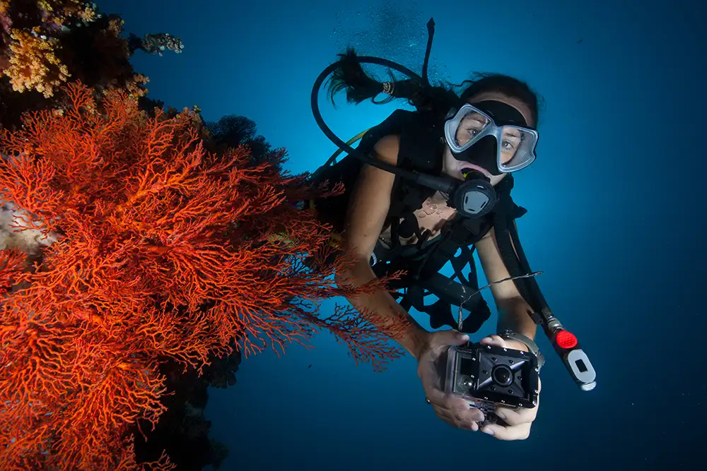 A lady in full scuba diving gear taking photos of coral reef with a gopro camera
