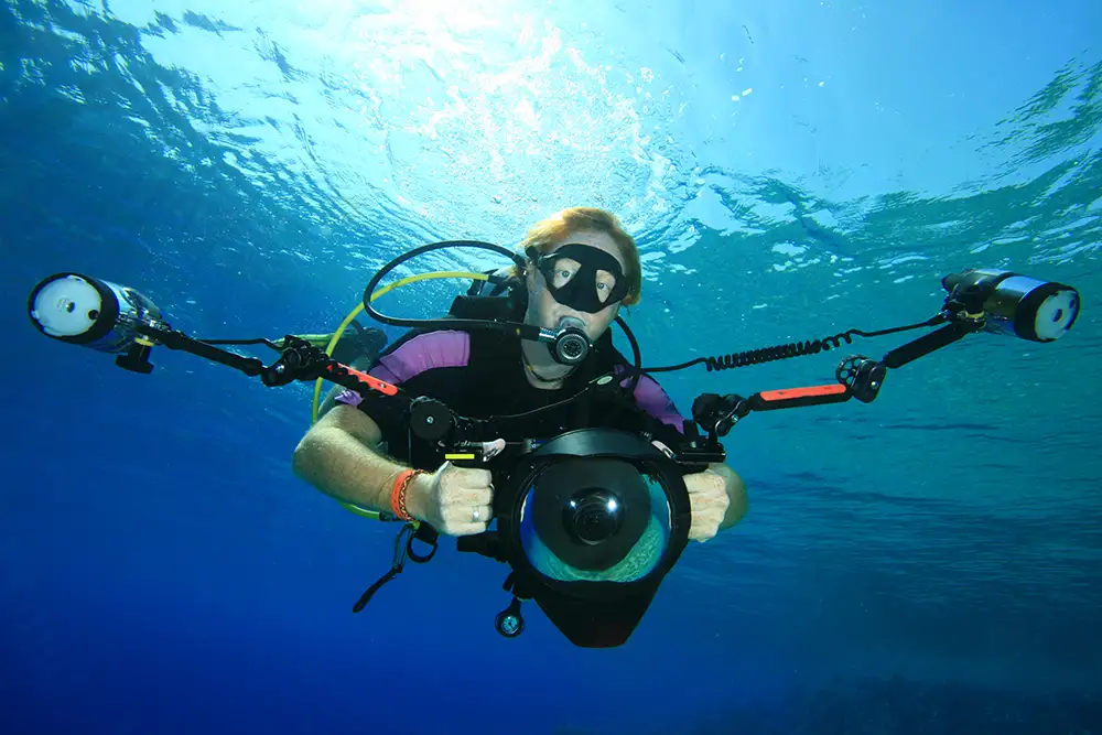 A man wearing full scuba diving gear underwater holding a large underwater camera with two lights either side