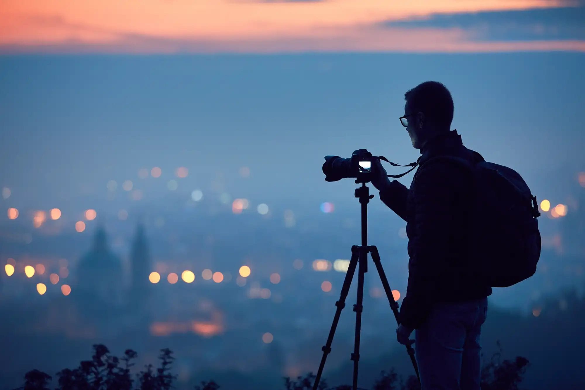 A silhouette of a photographer with a camera on tripod looking at a city in the distance at sunset